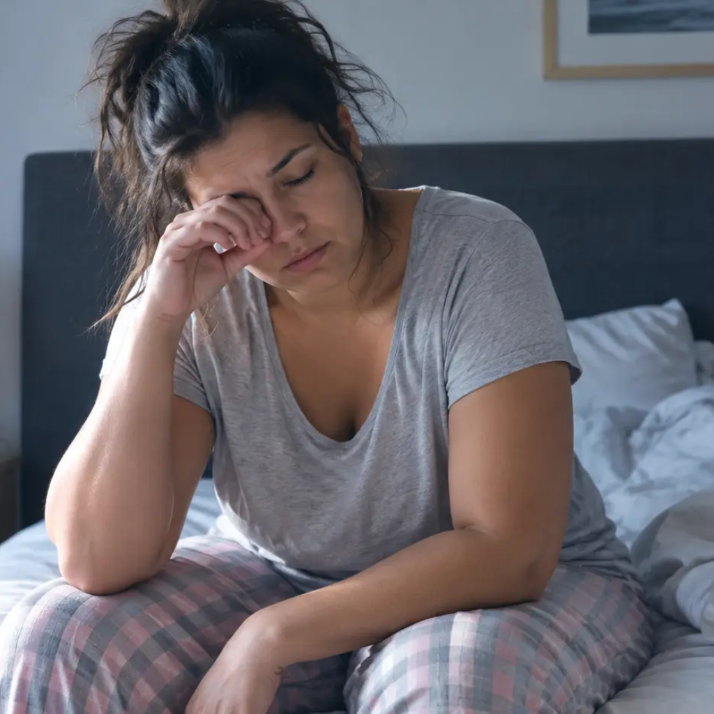 Femme fatiguée assise sur son lit au réveil, se frottant les yeux, illustrant la fatigue liée à l'apnée du sommeil silencieuse.